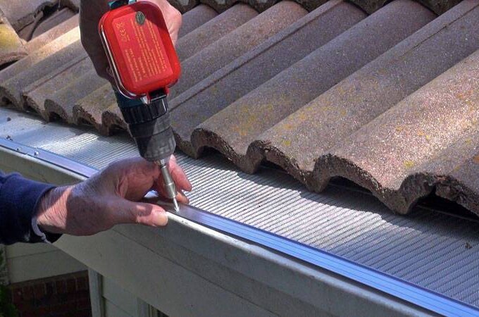 A person uses a power drill to install a stainless steel micro-mesh gutter guard along the edge of a tiled roof, securing it in place with a screw.