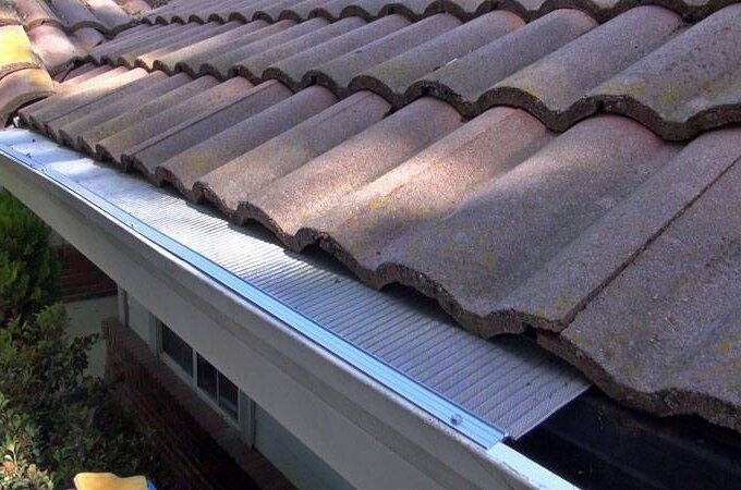 A close-up view of a house roof with curved clay tiles and a Gutterglove stainless steel micro-mesh gutter guard installed along the edge to prevent debris from entering the rain gutter below.