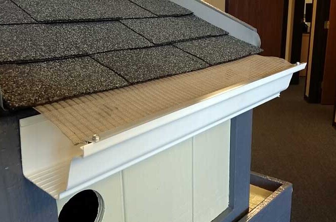 A close-up of a house roof with gray shingles and a white rain gutter fitted with a wire mesh gutter guard, designed to prevent debris from entering the gutter.