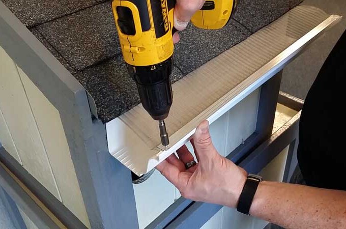 A person uses a yellow cordless drill to attach a white piece of trim to the edge of a small roof with gray shingles. One hand holds the trim in place while the other operates the drill.