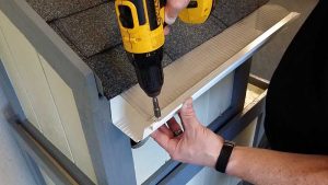 A person uses a yellow power drill to attach a white piece of trim to the edge of a small, shingled roof structure, holding the trim in place with one hand.