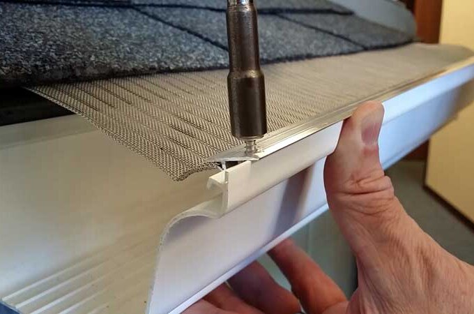 A person uses a drill to install a mesh gutter guard on a roof, securing it to the top edge of a white gutter under asphalt shingles.