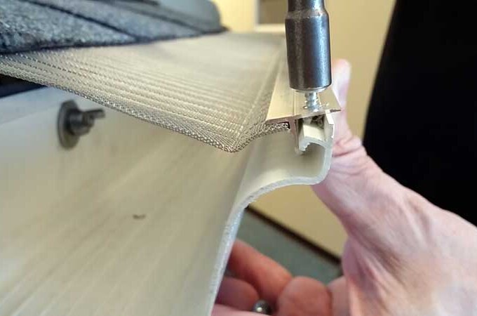 A close-up of a person’s hand holding an RV awning while a screwdriver tightens a screw into a metal bracket attached to the fabric.