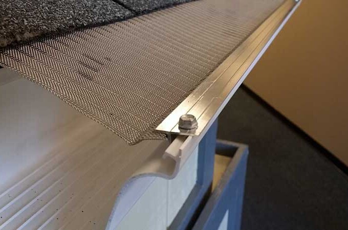 Close-up of a metal mesh gutter guard installed on the edge of a roof, showing the mesh secured with a metal strip and bolt to prevent debris from entering the gutter.