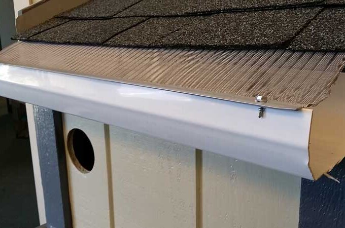 Close-up of a house roof edge with a white gutter and a metal mesh guard installed on top to prevent debris from entering the gutter. The roof has dark shingles and light-colored siding underneath.