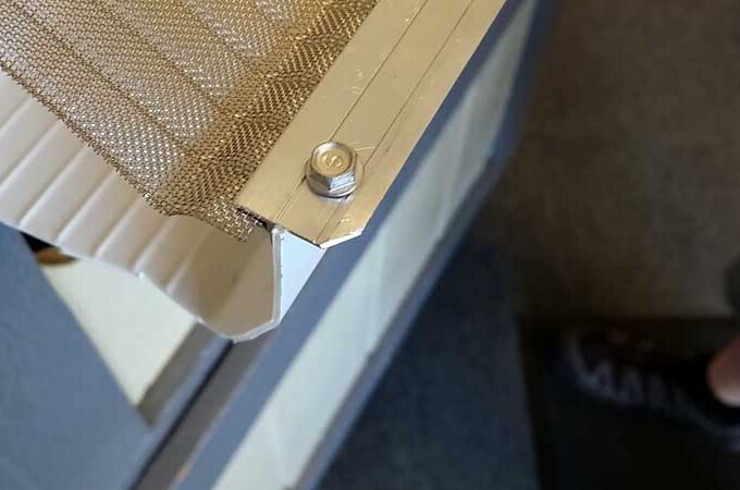 Close-up of a metal mesh attached to a corner frame with a bolt and nut, partially covering a corrugated white panel. A persons foot and leg are visible in the lower right corner on a carpeted floor.