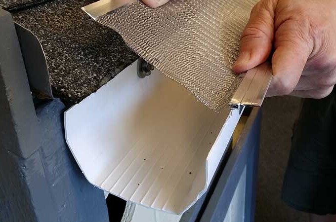A person installs a mesh gutter guard onto a house’s roof edge, positioning it above the gutter to prevent debris from entering. The hand holds the metal mesh sheet, aligning it over the white gutter.