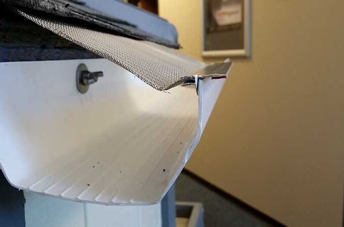 Close-up of a white rain gutter with a mesh guard partially installed, showing the metal screw attaching the gutter to a wall and an indoor hallway in the blurred background.