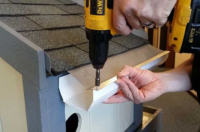 A person uses a yellow power drill to attach a white gutter to the roof edge of a small, light-colored birdhouse with gray shingles.