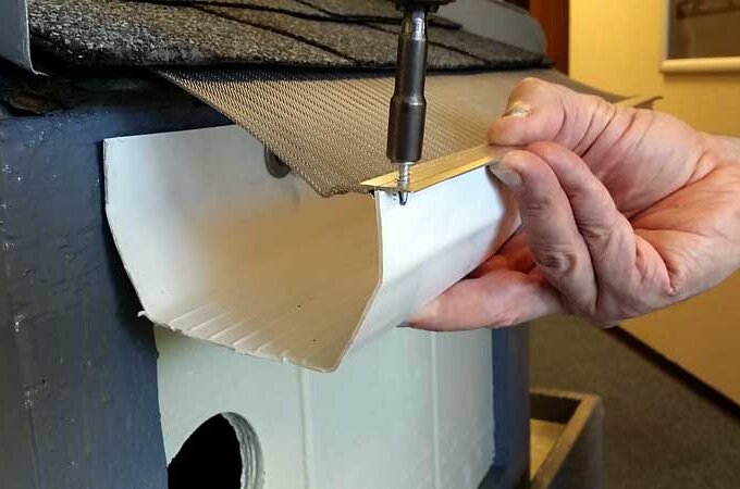 A person uses a screwdriver to attach a piece of metal mesh and a wood strip to the edge of a white gutter on a building.