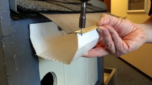 A person uses a power drill to attach a gutter guard mesh to the edge of a roof, securing it with a screw and metal bracket above a white rain gutter.