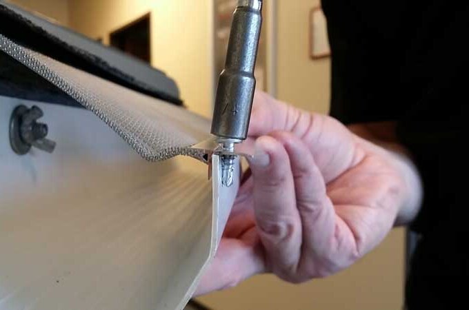 A person uses a drill to insert a screw through a metal strip and mesh material, attaching them to a white surface.