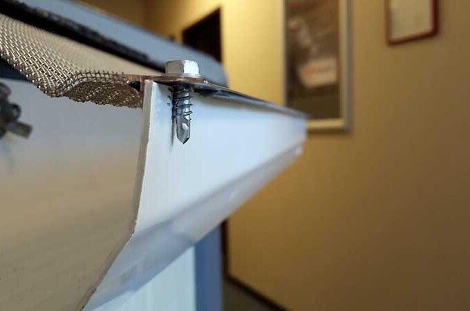 Close-up of a white rain gutter attached to a building, showing a metal mesh guard secured with a screw. The background is out of focus, revealing a hallway and framed items on the wall.