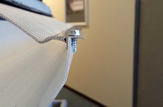 Close-up of a metal mesh screen attached to the edge of a white gutter with a screw, showing the installation detail. The background is out of focus.
