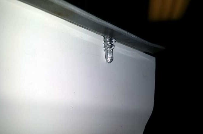 A close-up of a silver screw partially inserted into a white surface, with a metal edge above the screw and a dark, blurry background.