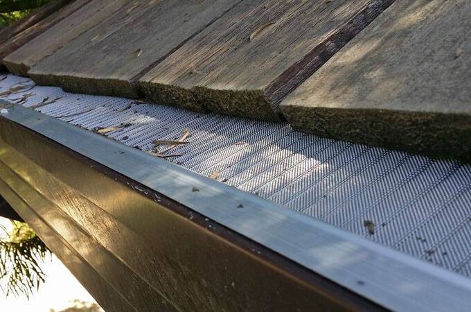 Close-up view of a house roof with a stainless steel micro-mesh gutter guard installed to prevent leaves and debris from clogging the gutter. Some small twigs and leaves rest on top of the guard, available at Costco.
