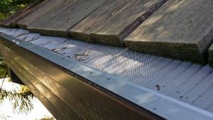 Close-up of a house roof with wooden shingles and a metal gutter guard installed, designed to prevent leaves and debris from entering the gutter. Some small debris is visible on top of the guard.