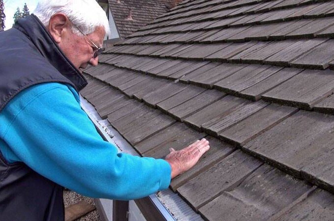 An older person wearing glasses and a blue jacket inspects the shingles on a sloped house roof, touching them with one hand while closely examining their condition, considering easy installation of stainless steel micro-mesh gutter guards.