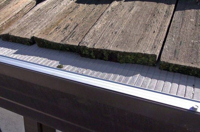 Close-up of a gutter with a stainless steel micro-mesh gutter guard installed beneath weathered, mossy wooden roof shingles, effectively preventing debris from entering the gutter.