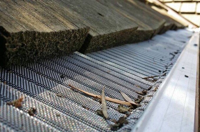 Close-up of a gutter with Gutterglove mesh guard installed under a wooden shingle roof, catching small debris like twigs and leaves. This solution from Costco offers easy installation for effective gutter protection.
