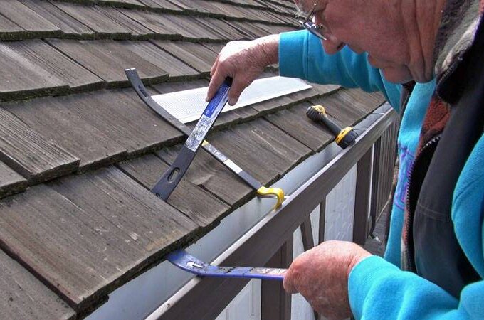 An older person uses tools to adjust or repair shingles on a house roof near the gutter, carefully working along the edge with a Gutterglove stainless steel micro-mesh.
