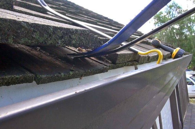 Close-up of a house roof with loose shingles being repaired; tools and cables are visible on the edge near the gutter guard, and trees, cars, and a driveway appear in the background.