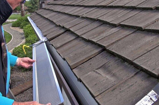 A person easily installs a stainless steel micro-mesh gutter guard along the edge of a roof with wooden shingles, preventing debris from entering the gutter. The surrounding yard is visible in the background.
