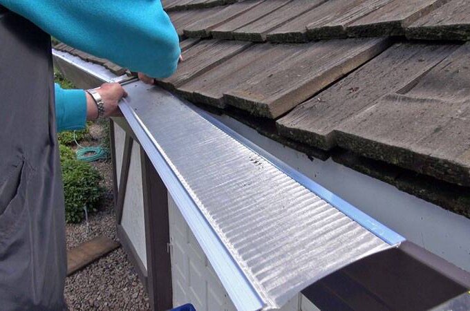A person installs an Easy On Gutter Guard along the edge of a sloped roof with wooden shingles, using a screwdriver to secure the gutter guard in place above a white building for easy installation.