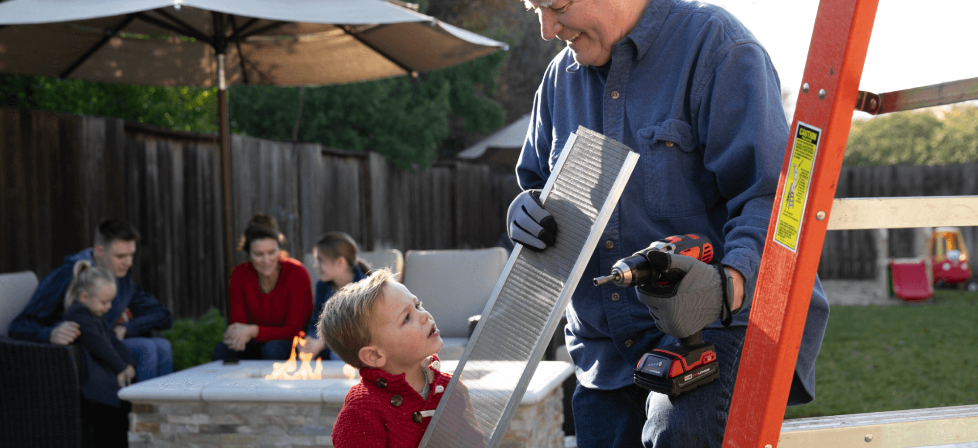 An older man holding a power drill and a metal vent works with a young boy beside a ladder in a backyard, possibly installing an Easy On Gutter Guard. In the background, a family sits around a firepit on patio furniture.