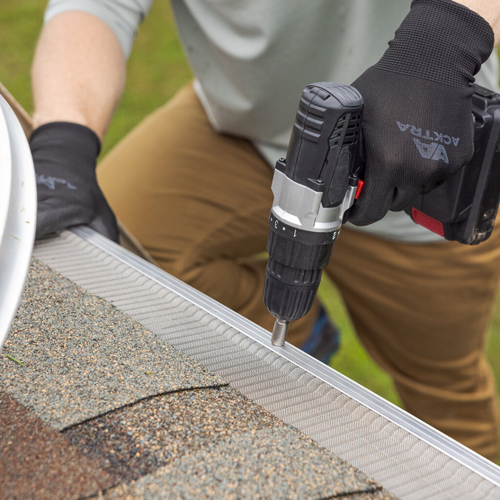 A person wearing gloves uses a cordless drill to install a metal gutter guard along the edge of a shingled roof. Grass can be seen in the background.